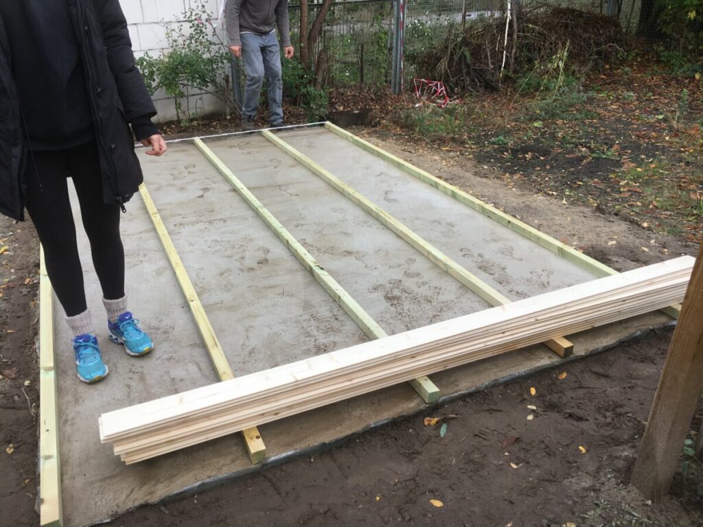 Auf das Fundament/die Bodenplatte wurden Holzleisten gelegt. Eine Person mit blauen Turnschuhen steht darauf.