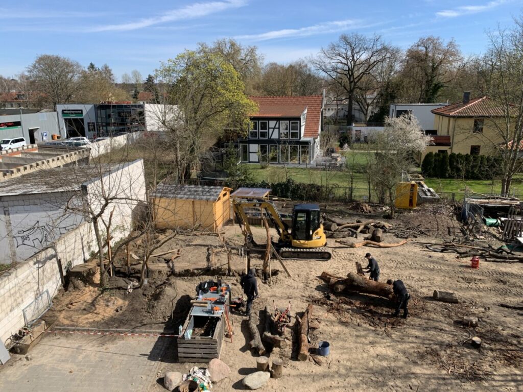 Luftbild des Schulhofs. Überwiegend sandiger Boden. Hinten das Gartenhäuschen, in der Mitte ein schmaler gelb-schwarzer Bagger, vorne drei Menschen.